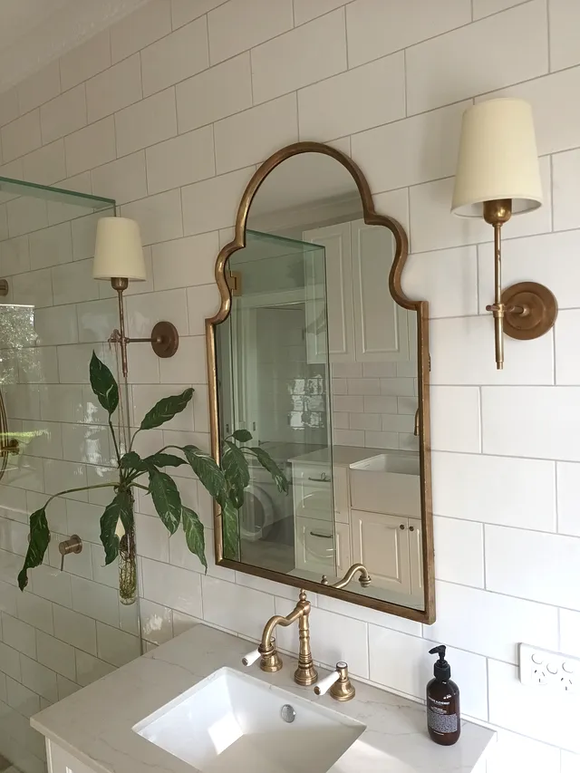 Arched brass mirror above a white shaker vanity against white subway tiles