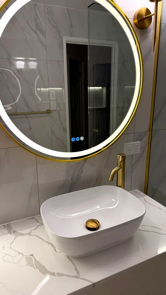 White vessel basin with brass tap and drain on a marble-look benchtop below a round gold LED mirror