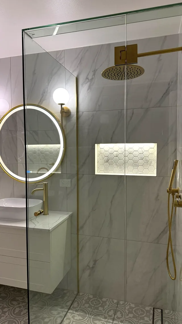 Frameless glass shower with a brushed brass rain head and a lit hexagon-mosaic niche