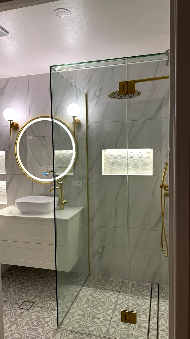 Bathroom with frameless glass shower, marble-look wall tiles, patterned floor tiles and a round LED mirror